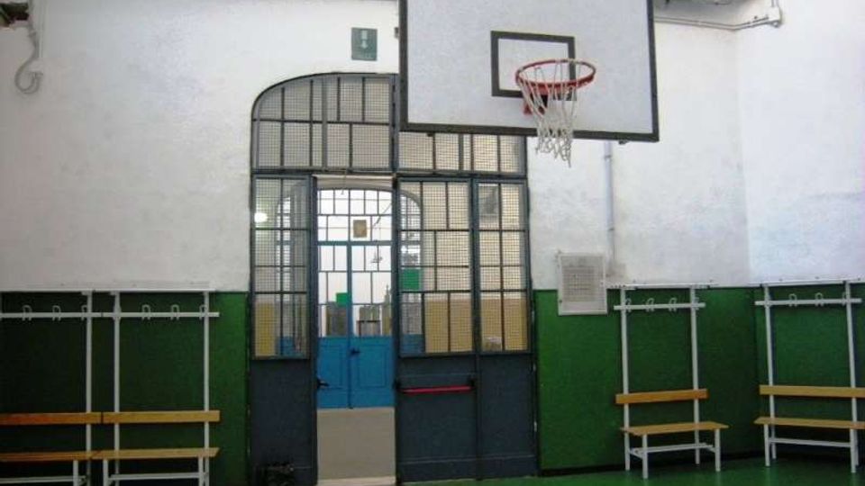 Interno di una palestra scolastica con pavimento verde, pareti bianche e verdi e canestro da basket montato sopra una porta ad arco con grate metalliche. Lungo le pareti sono presenti panche e appendiabiti.