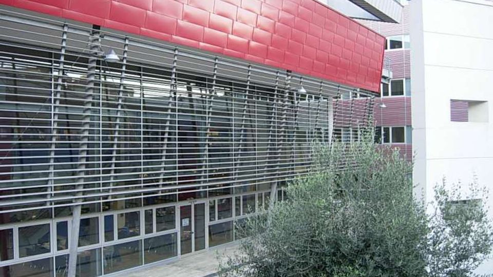 Facciata di un edificio moderno con rivestimento rosso a pannelli e ampia vetrata schermata da frangisole orizzontali in metallo, affacciata su un passaggio esterno. Sullo sfondo si vedono altri volumi bianchi e rosso scuro del complesso e alberi nell’area circostante.