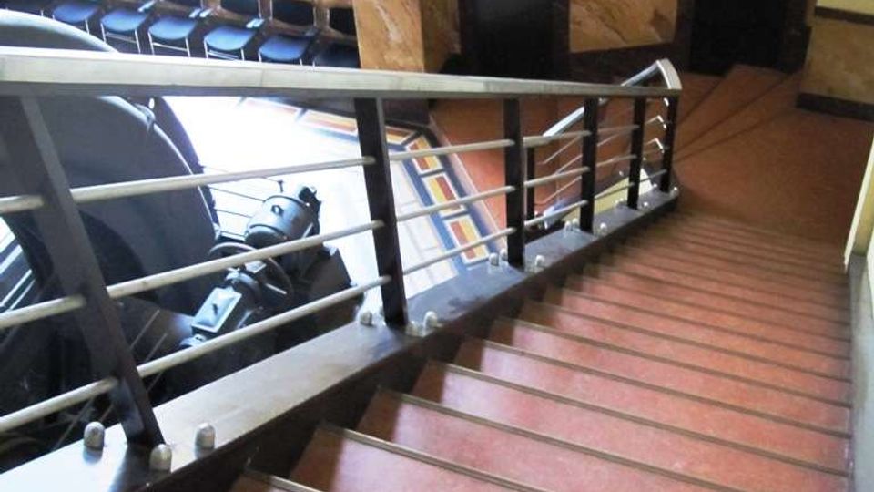 Scala interna in un edificio con gradini in rosso e corrimano in metallo, che scende verso un piano inferiore. Sullo sfondo si vedono una grande finestra a riquadri, pareti con rivestimento in marmo e due porte scure.
