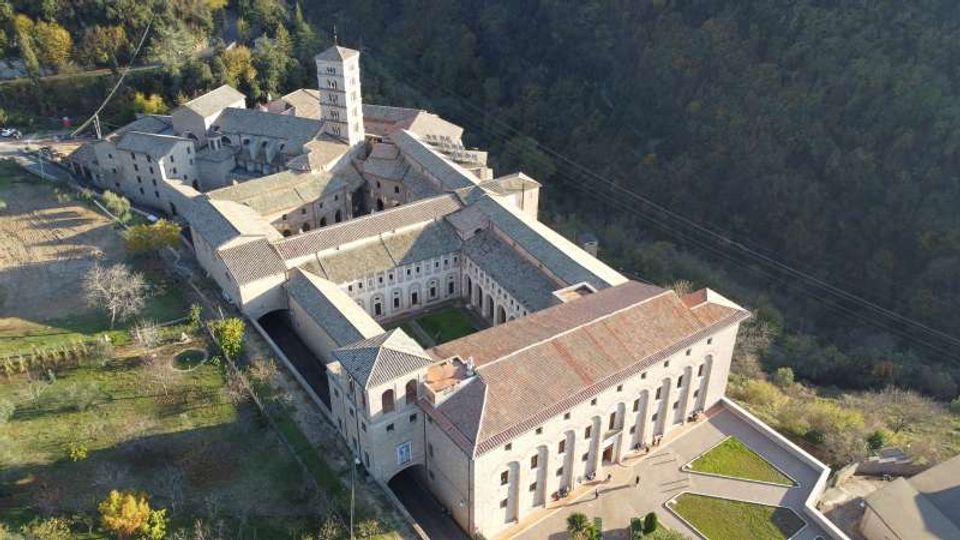 Veduta aerea di un grande complesso monastico in pietra con tetti in tegole, disposto attorno a un chiostro rettangolare e con un campanile quadrato centrale. L’edificio è circondato da aree verdi e da un bosco in collina, con cortili e vialetti lungo il perimetro.