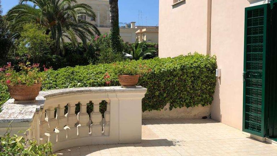Terrazza esterna pavimentata con balaustra in muratura e vasi di fiori, affiancata da una facciata color rosa con porta a grata verde. Sullo sfondo si vedono siepi e vegetazione con alte palme e altri edifici sotto cielo sereno.