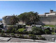 Veduta di una grande fortificazione in pietra con bastione arrotondato e mura alte, affacciata sul mare. In primo piano si vede un giardino urbano con palme, aiuole e vialetti, con una strada e alcuni autobus parcheggiati.