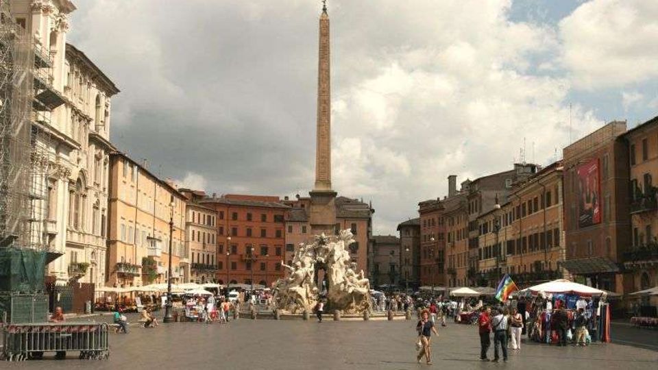 Ampia piazza urbana circondata da palazzi storici, con al centro una fontana monumentale con sculture e un alto obelisco. Ai lati della piazza si vedono edifici color ocra e arancio, aree con tende e bancarelle, sotto un cielo parzialmente nuvoloso.