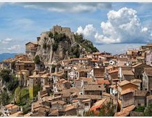 Veduta panoramica di un borgo storico con case addossate e tetti in tegole, disposto su un pendio roccioso. In alto domina una grande fortificazione in pietra costruita sulla rupe, con cielo parzialmente nuvoloso sullo sfondo.
