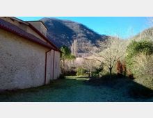 Lato esterno di un edificio in pietra con tetto in tegole rosse, affiancato da un prato e un sentiero erboso. Sullo sfondo si vedono alberi spogli e colline boscose ai piedi di una montagna sotto un cielo sereno.