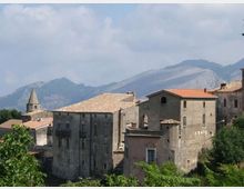 Complesso di edifici in pietra con tetti in coppi, tra cui una chiesa con campanile a guglia, affacciato su una valle verde. Sullo sfondo si vedono montagne e un cielo parzialmente nuvoloso.