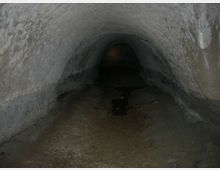 Interno di un tunnel sotterraneo con volta a botte in pietra grezza e pavimento umido. Il corridoio si restringe e scompare nell’oscurità verso il fondo.