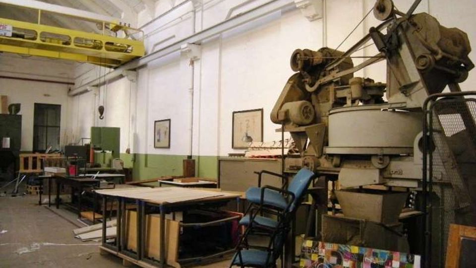 Interno di un capannone industriale con soffitto alto a volta, pareti bianche con fascia verde e una passerella carriponte gialla in alto. In primo piano si vedono grandi macchinari e attrezzature da officina, tavoli da lavoro e materiali accatastati lungo il lato destro.
