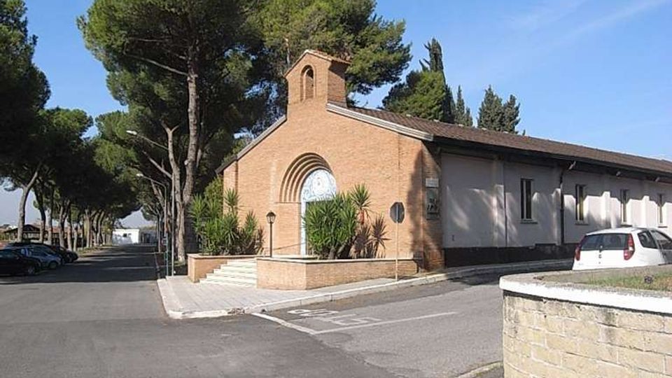 Piccola chiesa in mattoni con facciata semplice, arco sopra l’ingresso e campanile a vela, affiancata da un edificio basso e allungato. L’area circostante è una strada asfaltata con parcheggi e alberi alti (pini) lungo il viale, sotto un cielo sereno.