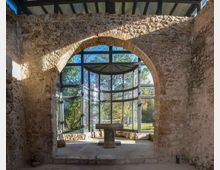 Interno di un edificio in pietra con grandi pareti grezze e un arco in muratura che incornicia un’abside moderna vetrata con struttura metallica. Oltre le vetrate si vede un’area verde alberata illuminata dalla luce naturale.