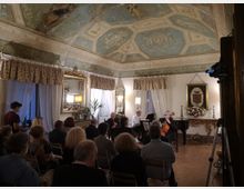 Sala storica adibita a concerto, con soffitto a volta decorato da affreschi e riquadri azzurri, pareti bianche con grandi finestre drappeggiate e specchi dorati. In fondo, un’area con pianoforte e arredi classici, tra cui una consolle intagliata con stemma incorniciato e composizioni floreali.