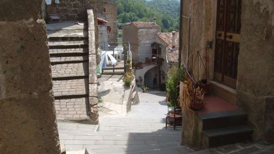 Vicolo in pietra in un borgo collinare, con scalinata e case in muratura ai lati. La strada scende verso un piccolo arco tra gli edifici, con tetti in coppi e colline boscose sullo sfondo.