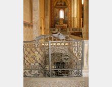 Interno di una chiesa con colonne in pietra e pareti affrescate; in primo piano una cancellata in ferro battuto decorata delimita un’area ribassata. Sullo sfondo si vede l’altare con tabernacolo e un’abside con affreschi e finestra centrale.