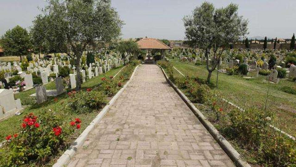 Viale pavimentato in un cimitero, fiancheggiato da aiuole fiorite e alberi, con file di lapidi e monumenti funerari su entrambi i lati. In fondo al percorso si vede una piccola struttura con tetto in tegole.