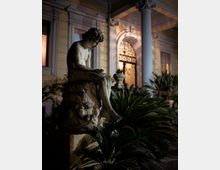 Statua in pietra su piedistallo in un giardino con palme nane e vegetazione, davanti alla facciata di una villa con colonne e finestre. Sullo sfondo si vede un portale ad arco con cancello decorato, illuminato in luce notturna.