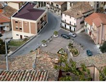 Veduta dall’alto di una piccola piazza di paese con parcheggio, aiuole e panchine, circondata da edifici a più piani con facciate chiare e tetti in tegole. Una strada curva costeggia la piazza e collega le case del centro storico.