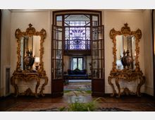 Interno di un elegante ingresso con porte a vetri aperte e grande finestra con vetrata decorata sopra l’architrave. Ai lati, due consolle e specchi barocchi dorati simmetrici; sullo sfondo si intravede un salotto con divano, tappeti e luce naturale dalle finestre.