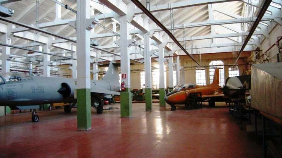 Ampio hangar industriale con pavimento rosso, colonne bianche e struttura del tetto a capriate con travi a vista. All’interno sono presenti diversi aerei e parti di velivoli, con grandi finestre sul fondo che illuminano lo spazio.