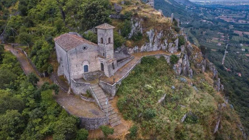 Chiesetta in pietra con campanile e tetto in tegole, costruita su un crinale roccioso con terrazze e una scalinata in muratura. Intorno ci sono boschi e pareti scoscese, con vista sulla valle e sui campi sottostanti.
