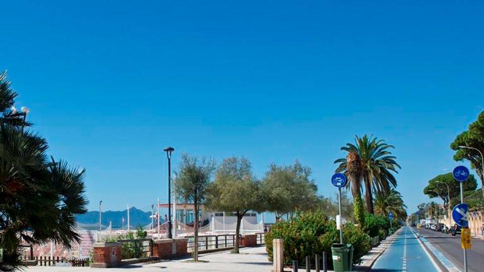Lungomare con ampia passeggiata pavimentata e pista ciclabile blu separata dalla strada, affiancate da palme, alberi e cartelli per biciclette. Sullo sfondo si intravedono la spiaggia e il mare con montagne lontane sotto un cielo sereno.