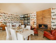 Sala da pranzo rustica con grande tavolo apparecchiato e sedie rivestite di bianco, pareti con carta da parati a cavalli e vetrina con bottiglie. A destra c’è un camino acceso incorniciato da pannelli di legno a spina di pesce, con legnaia sottostante e poltrone in pelle rossa.