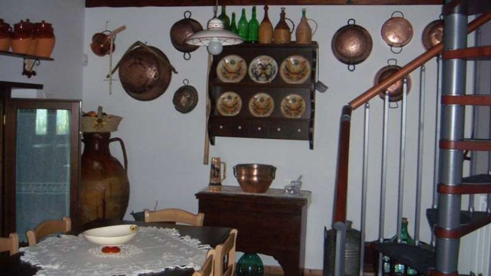 Interno rustico di una casa con soffitto a travi in legno, tavolo da pranzo con tovaglia e sedie, e pareti decorate con pentole di rame appese e una credenza con piatti e bottiglie. A destra si vede una scala a chiocciola metallica, con anfore e altri oggetti tradizionali disposti lungo le pareti.