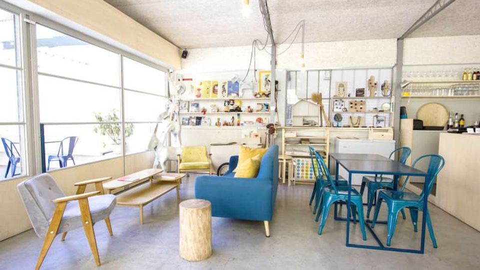 Interno luminoso di un loft con grandi vetrate scorrevoli su una terrazza, area salotto con divani e poltrone e tavolini bassi. Sul fondo si vede una cucina a vista con scaffalature e un tavolo da pranzo con sedie metalliche azzurre.