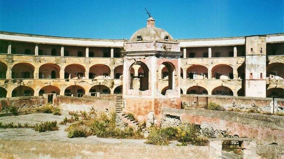 Cortile interno di una struttura storica in rovina, con un edificio semicircolare a due piani e una sequenza di arcate su entrambi i livelli. Al centro si vede un piccolo padiglione con cupola e portico, mentre il suolo del cortile è in parte spoglio e in parte invaso da vegetazione e detriti.