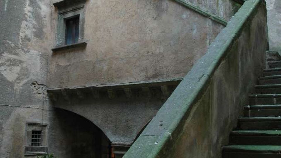 Cortile interno di un edificio storico con scala esterna in pietra che sale lungo un alto muro intonacato e in parte scrostato, con piccolo balcone e finestre. Alla base si vedono un arco di passaggio, pavimentazione in mattoni a spina di pesce e un vaso con pianta.
