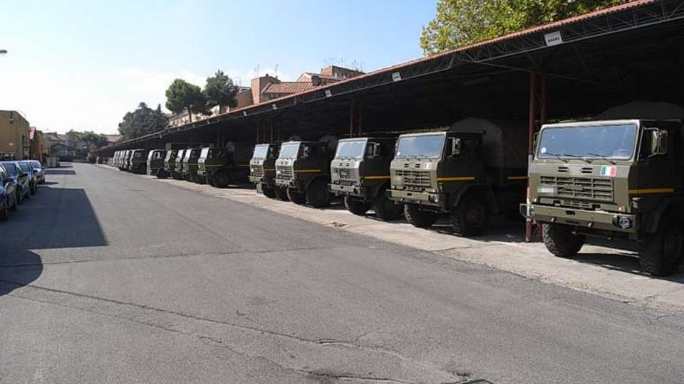 Ampio piazzale asfaltato in un deposito veicoli, con una lunga fila di camion militari parcheggiati sotto una tettoia metallica con copertura rossa. Sullo sfondo si vedono edifici e alberi lungo il perimetro dell’area.