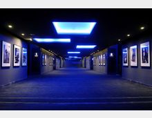 Corridoio interno moderno e ampio, con pareti scure rivestite da fotografie incorniciate ai lati e pavimento in moquette. Il soffitto presenta grandi pannelli luminosi rettangolari con illuminazione blu, creando una prospettiva centrale verso il fondo del passaggio.