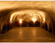 Interno di una grotta o cantina sotterranea con soffitto a volta scavato nella roccia e pavimento in cemento. Luci calde lungo le pareti illuminano una serie di nicchie e archi sul fondo.