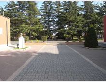 Piazzale pedonale pavimentato con blocchetti e corsie laterali in pietra, affiancato da siepi potate e lampioni. Sullo sfondo si vedono alti alberi sempreverdi e, a lato, edifici con facciate semplici e una piccola statua su piedistallo.