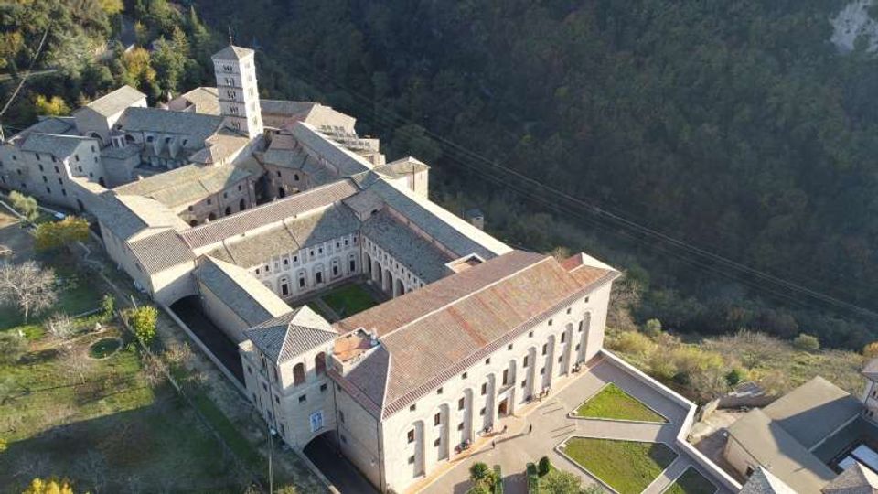Veduta aerea di un grande complesso monastico in pietra con tetti in coppi e una torre campanaria, organizzato attorno a un chiostro interno. L’edificio sorge ai margini di un’area boscosa su un pendio, con cortili e spazi verdi perimetrali.