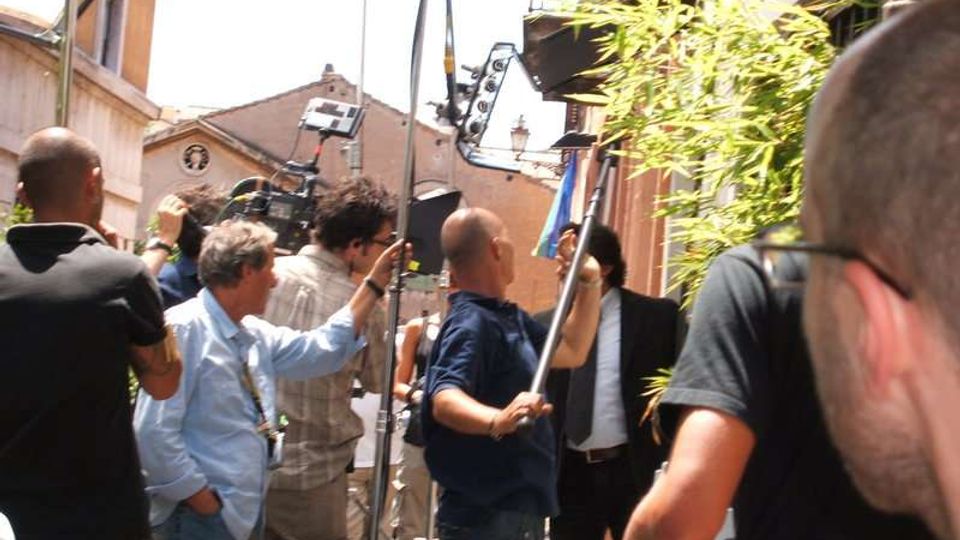 Ripresa in una stretta via urbana con edifici bassi e facciate in intonaco, attrezzature da set cinematografico come telecamera su supporto, aste e pannelli di illuminazione. Sul lato destro si vede una fitta pianta di bambù davanti a un edificio.