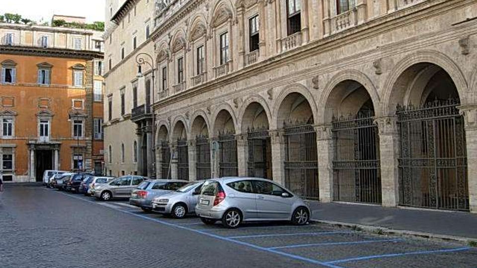 Strada urbana lastricata con auto parcheggiate lungo il marciapiede, affiancata da un grande edificio storico in pietra con porticato ad archi e cancellate in ferro. Sullo sfondo si vedono altri palazzi in stile classico con facciate ocra e finestre ornate.
