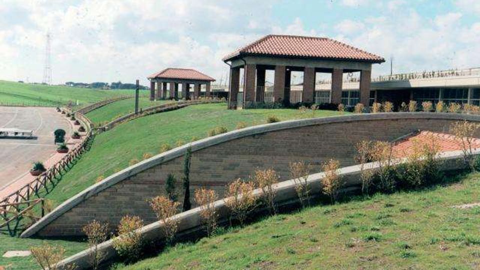 Area verde in collina con un muro curvo di contenimento in mattoni e una passerella sopraelevata. In cima si vedono due piccoli padiglioni aperti con tetto in tegole e, sullo sfondo, un ponte o viadotto.