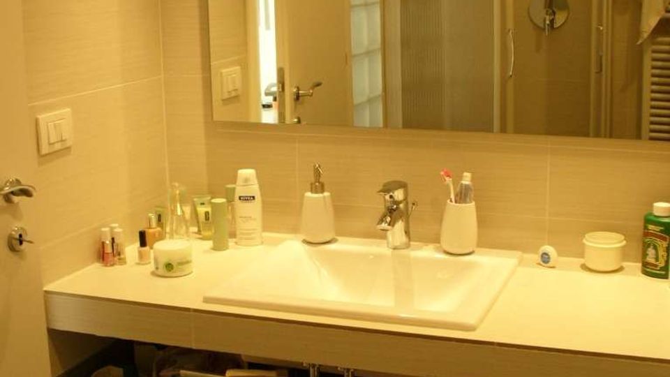Bagno moderno con lavabo rettangolare su mobile sospeso e grande specchio a parete; sul piano sono disposti vari prodotti da toilette. Sullo sfondo si vede una cabina doccia con porte scorrevoli e pareti piastrellate in tonalità chiare.
