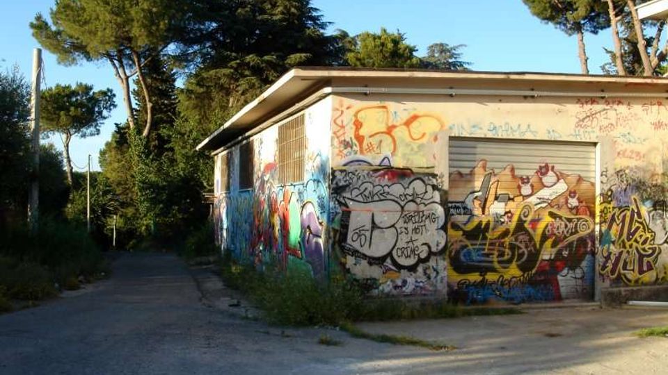 Piccolo edificio basso in cemento con tetto piatto e una serranda metallica, le pareti completamente coperte da graffiti e murales. La struttura si trova ai margini di una strada asfaltata, circondata da alberi alti e vegetazione.
