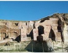 Rovine di un complesso in mattoni incassato in una parete di tufo, con grandi arcate e nicchie scavate nella roccia. In alto si vede il bordo del pendio con una ringhiera, sotto un cielo azzurro.