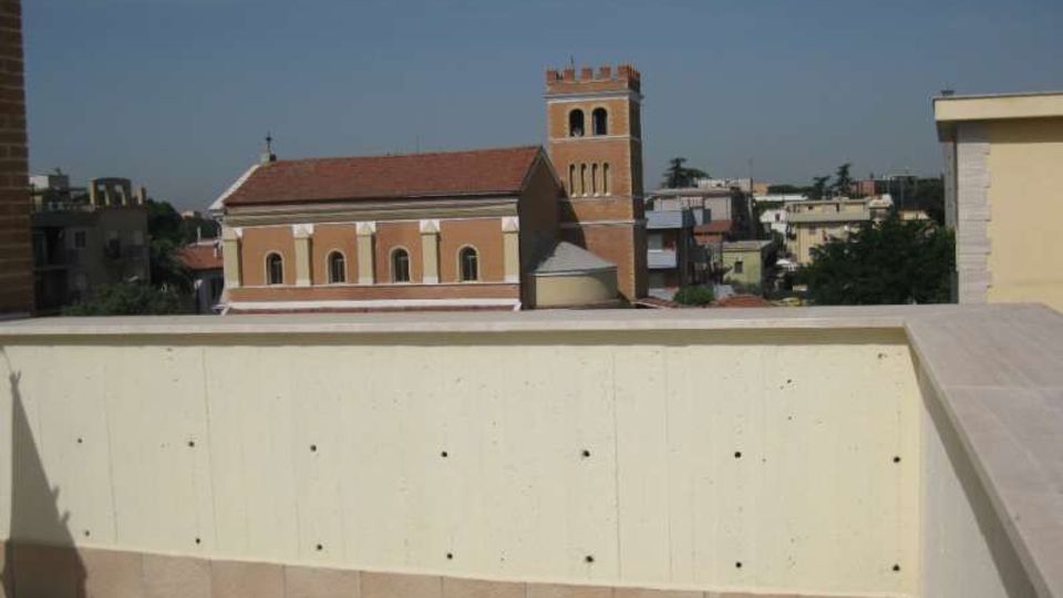 Terrazza con pavimentazione in piastrelle e parapetto in muratura chiara, con vista su una chiesa in mattoni dal tetto rosso e campanile merlato. Sullo sfondo si vedono edifici residenziali e alberi sotto un cielo sereno.