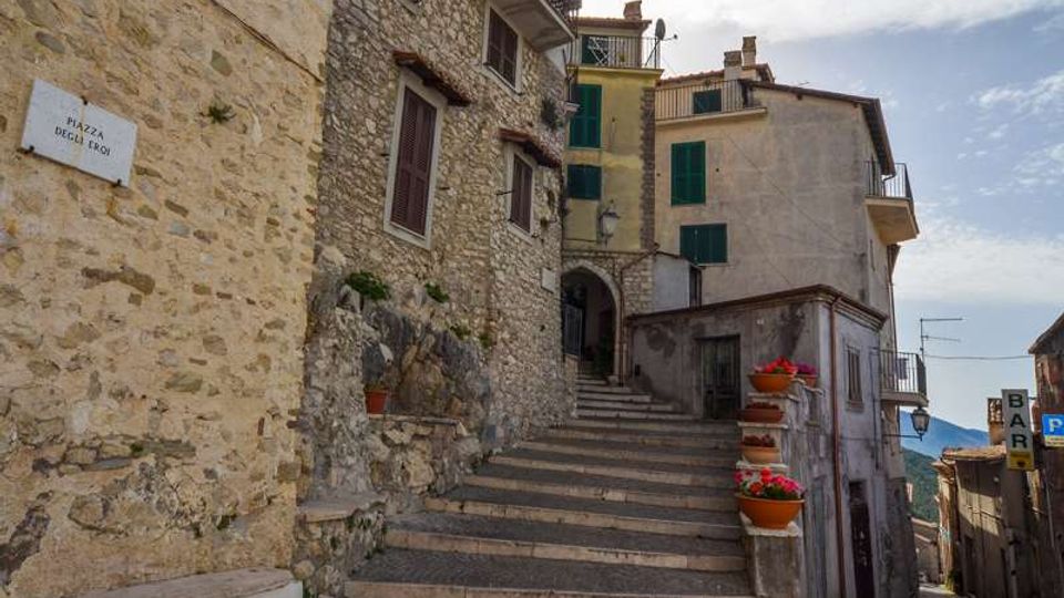 Scalinata in pietra in un borgo storico, affiancata da case in muratura con persiane verdi e balconi. Lungo i gradini ci sono fioriere con fiori rossi, con una stretta via che scende sulla destra e un’insegna “BAR” visibile sullo sfondo.