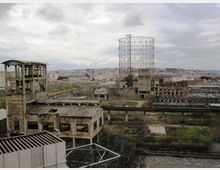 Vista panoramica di un complesso industriale dismesso con capannoni in muratura e strutture metalliche deteriorate, dominato da un grande gasometro cilindrico in traliccio. Sullo sfondo si vede la città sotto un cielo nuvoloso, con aree verdi e binari o passerelle sopraelevate nell’area.