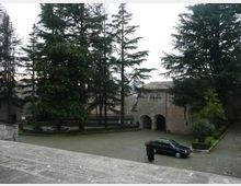 Cortile in pietra con vialetti e aiuole, circondato da alti alberi sempreverdi; sullo sfondo un edificio storico in mattoni con portico ad archi e finestre ad arco. In primo piano si vedono gradini in pietra che scendono verso lo spazio aperto.