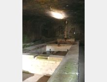 Interno in penombra di un antico lavatoio in muratura, con vasche rettangolari in pietra e acqua stagnante. La struttura è coperta da una volta bassa e presenta pareti consumate dall’umidità e un rubinetto centrale.
