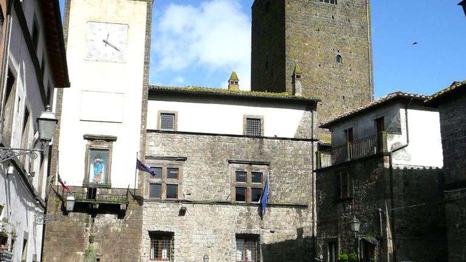 Piazza lastricata in un borgo storico con edifici in pietra: in primo piano un piccolo parcheggio e, sullo sfondo, una torre campanaria con orologio e un’alta torre medievale. Facciate con finestre a grata, balconi e tetti in coppi sotto un cielo azzurro.