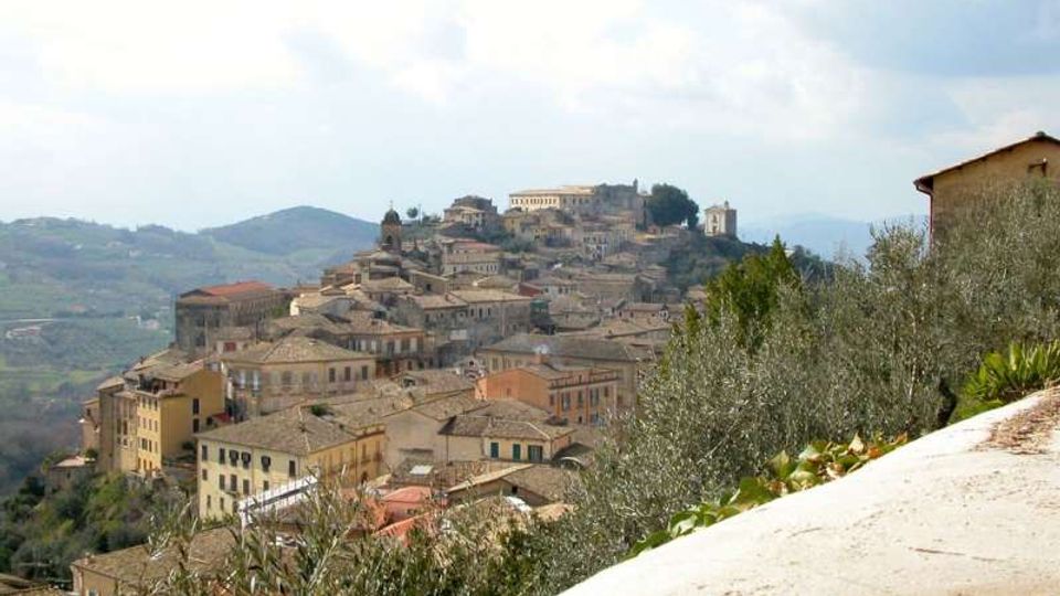 Panorama di un borgo collinare con case in pietra dai tetti in coppi, disposto a gradoni sul pendio e dominato da edifici storici e una torre campanaria. In primo piano si vedono vegetazione e un muretto, con colline e montagne sullo sfondo sotto un cielo nuvoloso.