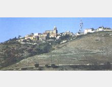 Panorama di un piccolo borgo su un crinale collinare, con case compatte e una chiesa con campanile al centro. In primo piano si vede un pendio brullo con tracce di sentieri e, sulla destra, un’alta antenna di telecomunicazioni.