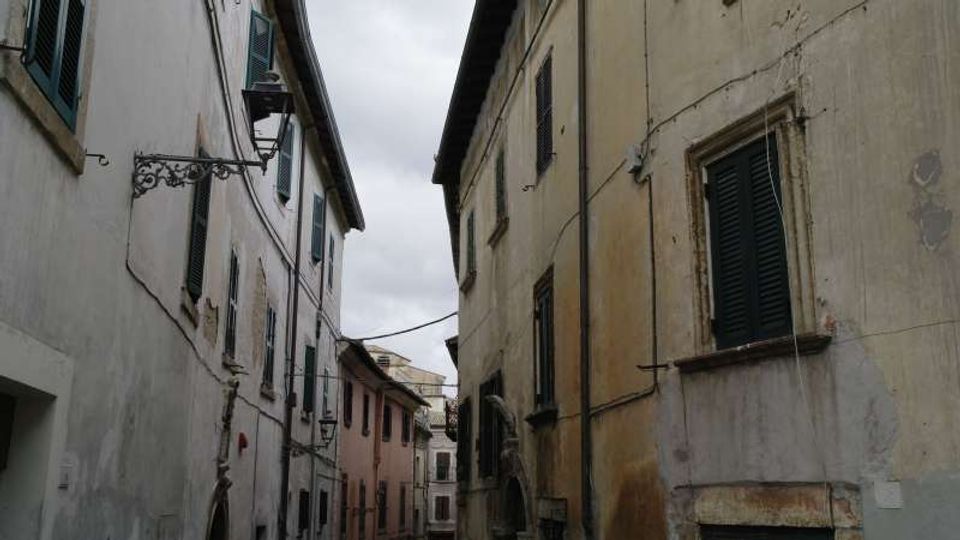 Stretta via di un centro storico tra edifici alti in muratura con facciate intonacate e persiane scure, illuminate da lanterne a muro. Cavi sospesi attraversano la strada sotto un cielo nuvoloso.