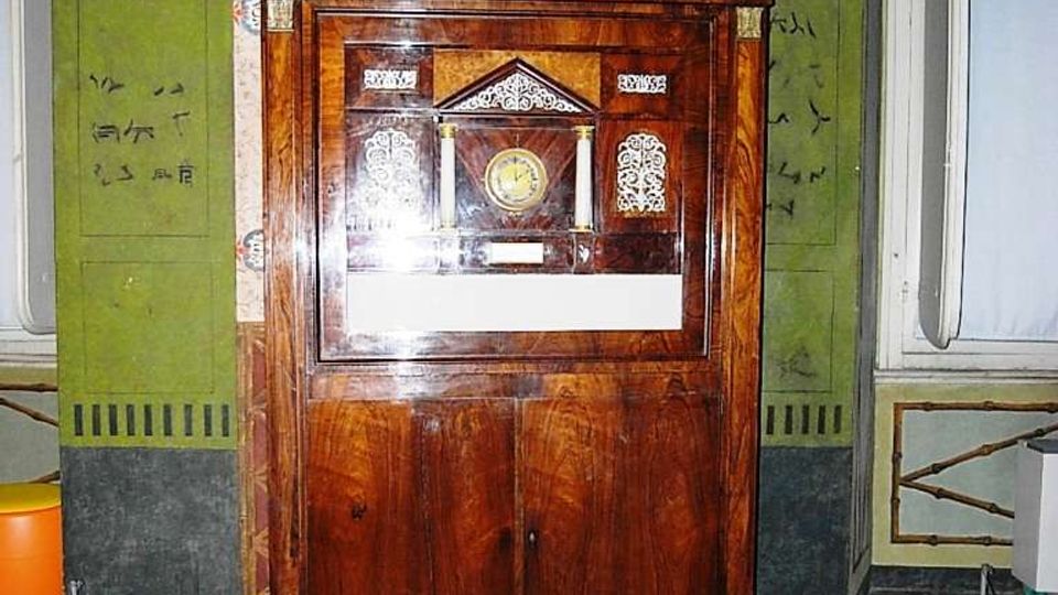 Mobile antico in legno lucido con vetrina, simile a una pendola o a un espositore con quadrante e decorazioni interne, collocato su una pedana e circondato da cordoni di protezione. Sullo sfondo si vede una parete decorata con pannelli verdi con scritte e motivi ornamentali e un pavimento in parquet.